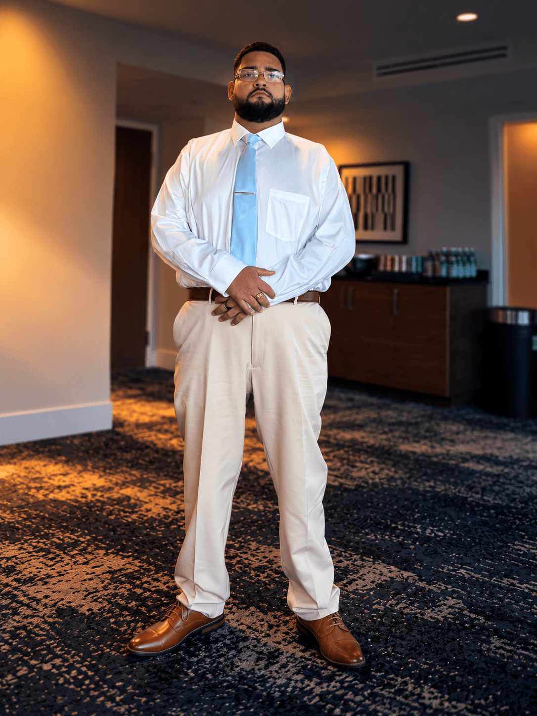 Emilio Sierra standing indoors in a white shirt, light blue tie and beige trousers.